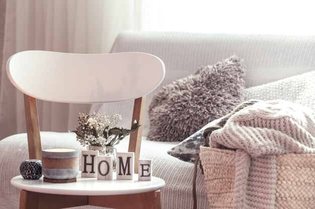 Chic interior for a house. Candles, a vase with flowers with wooden letters of the home on wooden white chair. Sofa and wicker basket with cushions in the background. Home decoration.