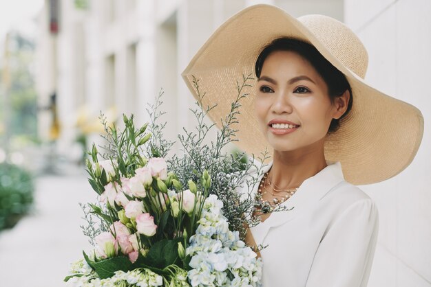 신선한 꽃 부케와 거리에서 포즈를 취하는 큰 밀짚 모자에 세련된 부유 한 아시아 여자