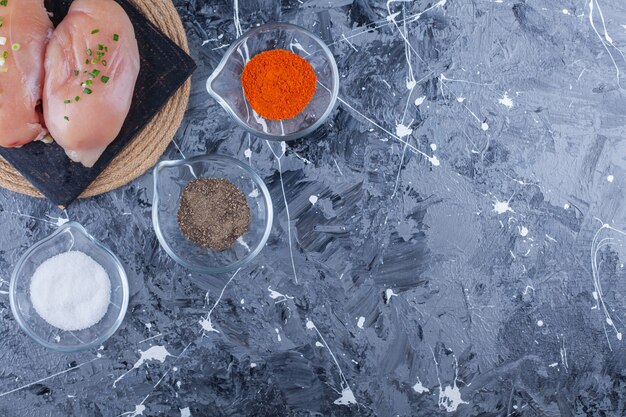 Chicken breast on a cutting board on a trivet next to bowls full of spices, on the blue table.