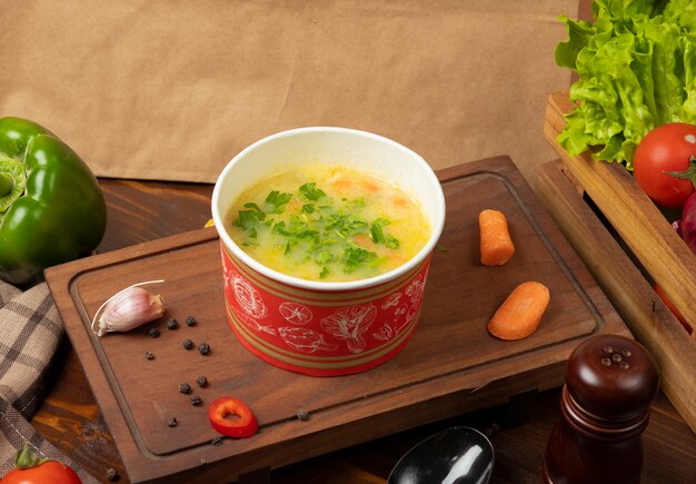 Chicken broth vegetable soup in disposable cup bowl served with green vegetables.