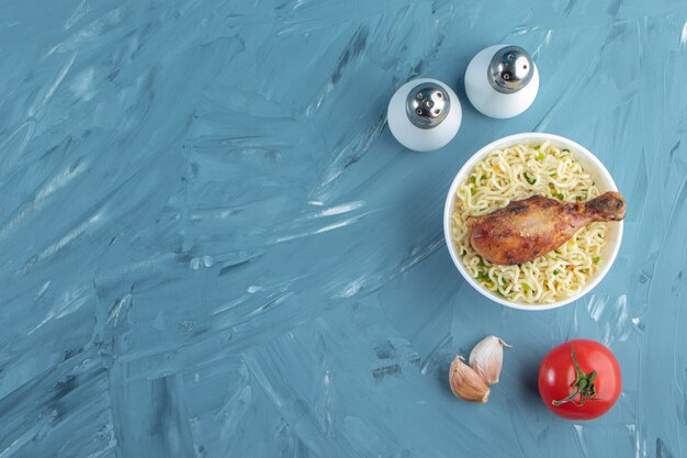 Chicken drumsticks and noodle in a bowl next to salt, tomatoes and garlic, on the marble background. 