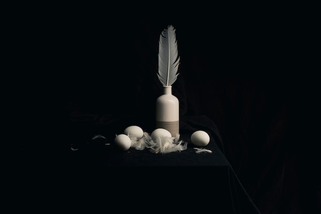 Chicken eggs with feathers near quill in vase on edge of table between blackness