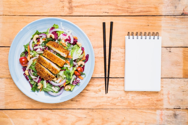 Куриное филе с салатом на керамической тарелке; палочки для еды и пустой спиральный блокнот на деревянном столе