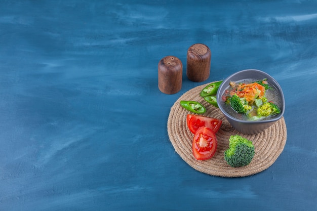 Chicken soup in a bowl and sliced vegetables on a trivet, on the blue table.