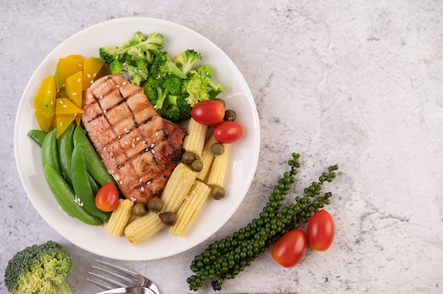 Bistecca di pollo condita con sesamo bianco, piselli, pomodori, broccoli e zucca in un piatto bianco.