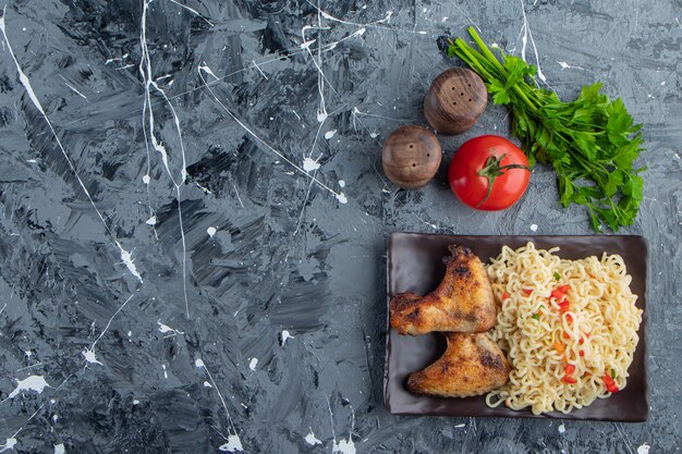 Куриные крылышки и лапша на блюде рядом с овощами на мраморном фоне.