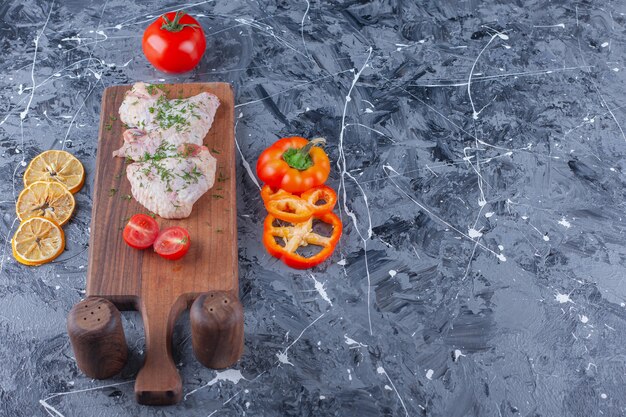 Chicken wings and sliced tomatoes on a cutting board next to assorted vegetables, on the blue background. 