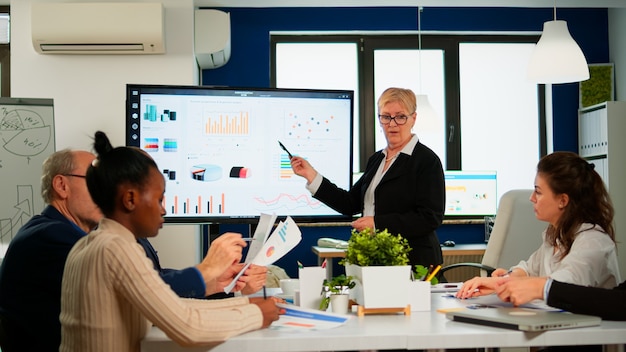 Foto gratuita analista capo che tiene la presentazione della riunione per un team di economisti. manager che mostra lavagna interattiva digitale con analisi della crescita, grafici, statistiche, dati, diverse persone che lavorano in broadroom