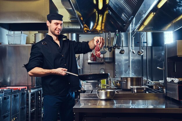 The chief cook holds pork meat under the fry pan in a kitchen.