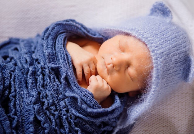 Child in blue hat and knitted scarf sleeps on white pillow 