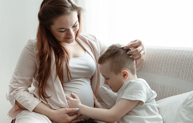Ребенок мальчик обнимает живот беременной ее матери на диване у себя дома.