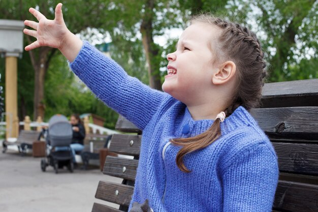 Child girl in a blue knitted sweater eats ice cream sitting on a bench.