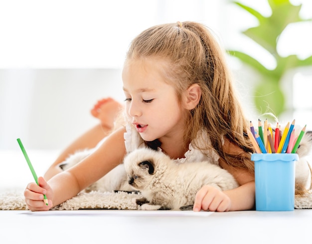 Ребенок девочка рисует с котятами тряпичной куклы и лежит на полу. маленькая женщина рисует красочными карандашами и домашних животных котенка рядом с ней дома
