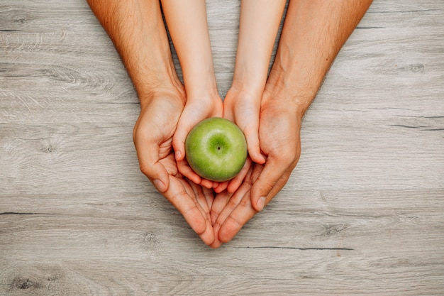 Free photo child holding a green apple in her hands inside of her father's hands top view on a light wooden