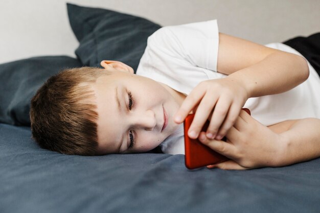 Ребенок лежит на кровати и использует вид сбоку мобильного телефона