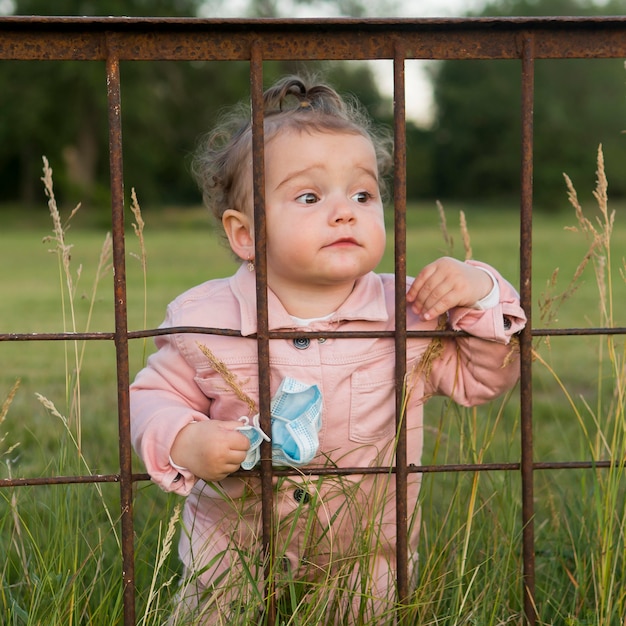 Child in pink clothes behind park bars holding medical mask