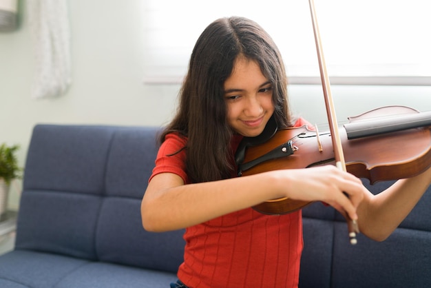 バイオリンを弾く子供。自宅で音楽のレッスンを練習しているラテン系の女の子の笑顔。新しい楽器を演奏することを学ぶためにオンラインクラスを受講している若い学生