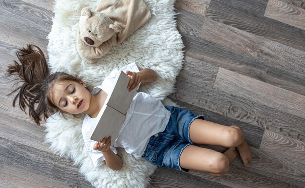 The child reads a book lying on a cozy rug at home with his favorite toy teddy bear.