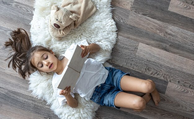 Ребенок читает книгу, лежа на уютном коврике дома со своим любимым игрушечным мишкой.