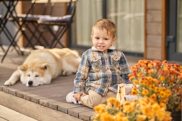 아이, 안전. 격자 무늬 셔츠와 베이지색 바지를 입은 귀여운 웃는 아이가 꽃 근처 집 베란다에 앉아 있고 좋은 날에 누워 있는 개를 바라보는 카메라를 보고 있습니다.