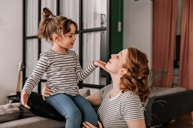 Child in striped T-shirt is sitting on sofa and stroking his mothers face.