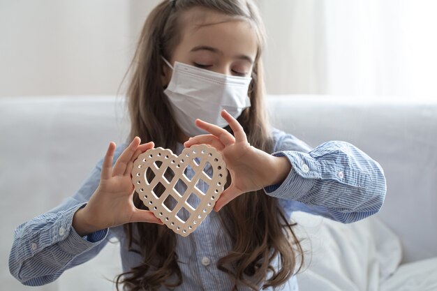 Child wearing medical protective face mask to health protection from coronavirus, holds wooden heart.