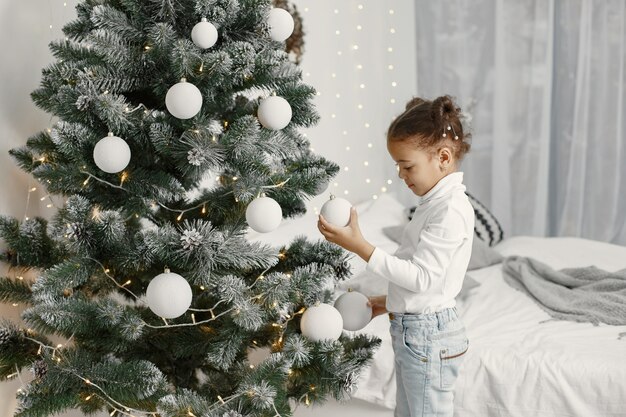 Child in a white sweater. Daughter standing near Christmas tree.