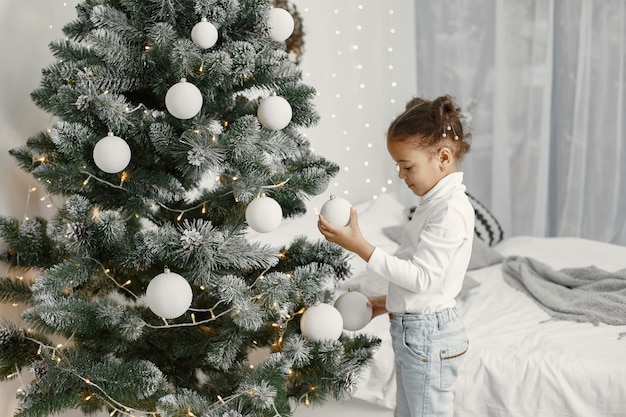 Child in a white sweater. Daughter standing near Christmas tree.