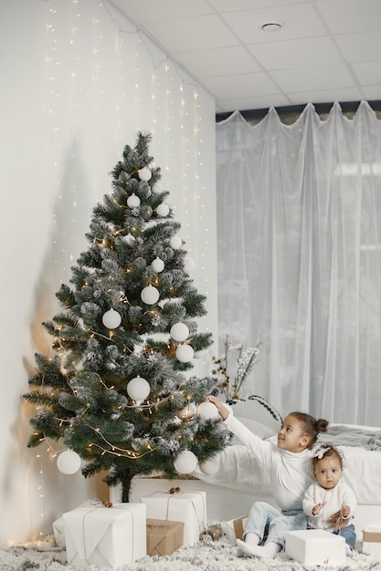 Child in a white sweater. Daughters stitting near Christmas tree.Two sisters at home.