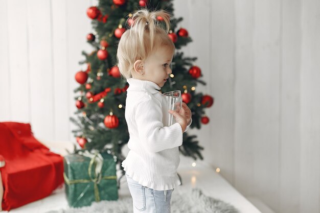 Child in a white sweater is playing. Daughter is standing near Christmas tree.