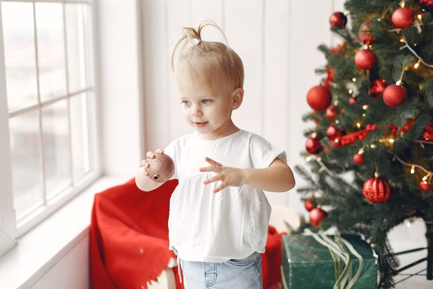 白いTシャツを着た子供が遊んでいます。クリスマスツリーの近くに座っている娘。