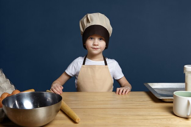 Childhood, hobby, food, nutrition, cooking, baking and bakery concept. Isolated shot of excited handsome blue eyed Caucasian little boy wearing chef uniform making pastry in the kitchen, copy space