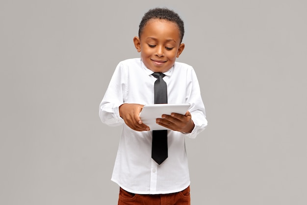 Детство, современные технологии и вредные привычки. Симпатичный афроамериканский школьник, пристрастившийся к электронным гаджетам, использует цифровой планшет, чтобы играть в видеоигры, впитав выражение лица