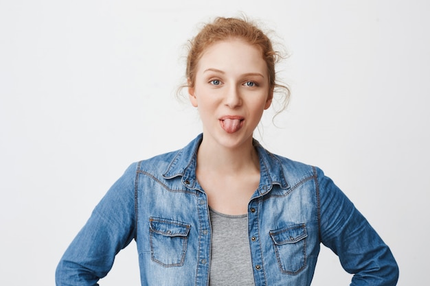 Childish emotive redhead teenage girl sticking out tongue and smiling, make faces while standing with hands on waist