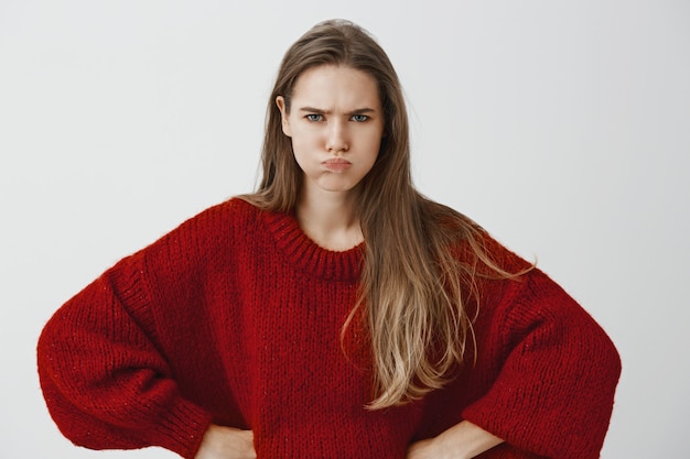 Childish girlfriend wants attention. portrait of displeased offended european woman in red loose sweater, holding hands on hips, sulking and frowning, arguing and being insulted