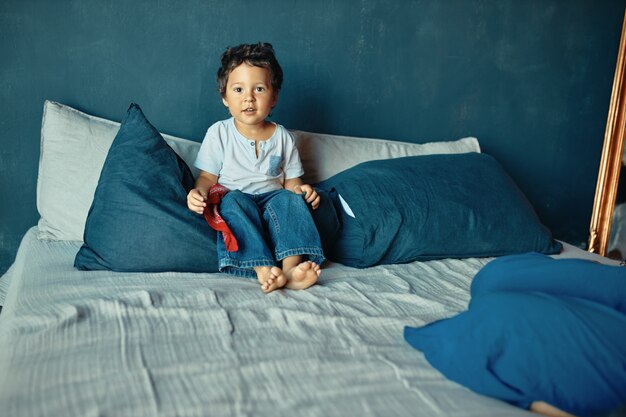Дети, постельные принадлежности и концепция воспитания. Милый босиком смешанной расы маленький мальчик сидит на кровати, готовый играть после дневного сна.