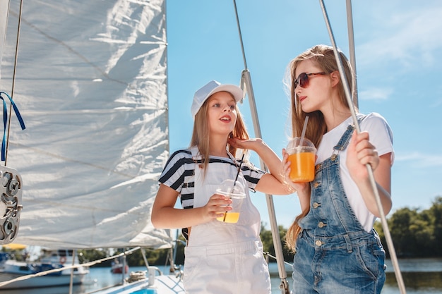 オレンジジュースを飲む海のヨットに乗っている子供たち。屋外の青い空に対して10代または子供の女の子。カラフルな服。キッズファッション、晴れた夏、川、休日のコンセプト。