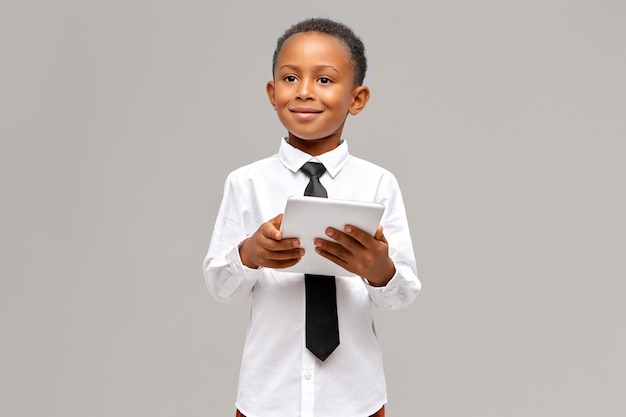 Children, electronic devices and gadgets concept. Smart confident African pupil in uniform posing isolated with portable touch pad computer in his hands, surfing internet or shopping online