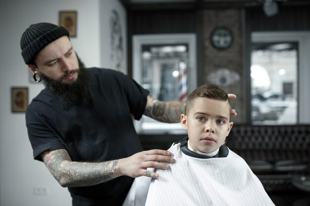 Детский парикмахер резки маленького мальчика на темном фоне. Довольный милый мальчик дошкольного возраста стрижка. На руке мастера татуировка со словом бритье