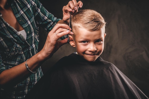 Children hairdresser with scissors is cutting little boy against a dark background. Contented cute preschooler boy getting haircut.