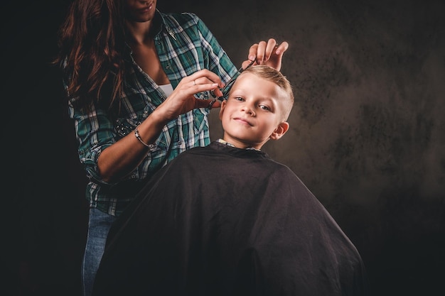 はさみを持つ子供の美容師は、暗い背景に対して小さな男の子をカットしています。ヘアカットを取得している満足のいくかわいい未就学児の男の子。