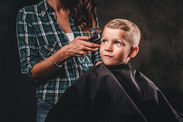 トリマーを持つ子供の美容師は、暗い背景に対して小さな男の子をカットしています。かわいい未就学児の男の子が散髪をしています。