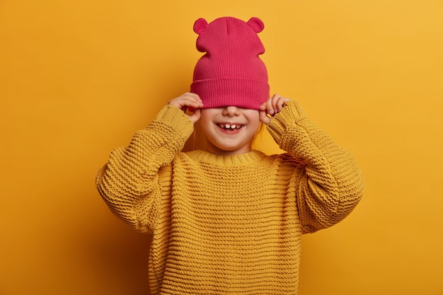 Children, happiness, wellbeing concept. Carefree playful girl covers half of face with hat, tries to hide from someone, wears loose knitted sweater, isolated on yellow wall, got pretty smile