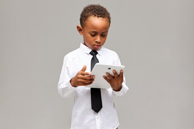 Children and modern technology concept. Serious focused Afro American schoolboy in uniform holding white generic digital tablet, playing online game or learning, having concentrated expression