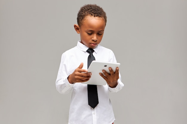 Дети и концепция современных технологий. Серьезный сосредоточенный афроамериканский школьник в униформе держит белый цифровой планшет, играет в онлайн-игру или учится, сосредоточенно выражая свои мысли