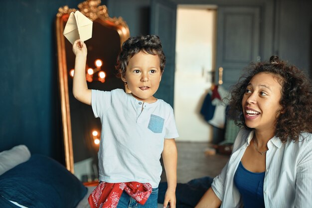 Children, motherhood, fun and hobby concept. Indoor Portrait of excited emotional dark skinned little boy standing on bed with hand up