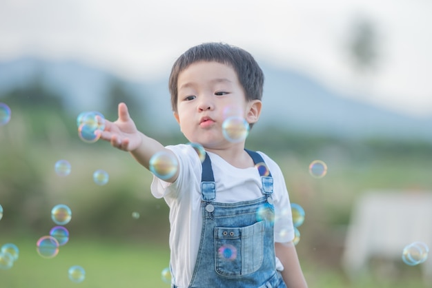 こどもの日。公園でシャボン玉を吹く少年。夏の畑でシャボン玉で遊ぶかわいい幼児の男の子。手を挙げろ。幸せな子供時代のコンセプト。本格的なライフスタイルイメージ。