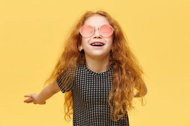Дети, стиль и концепция fshion. Беззаботная модная маленькая девочка с вьющимися рыжими волосами, со счастливым радостным выражением лица, смеющаяся, в стильных розовых солнцезащитных очках и держащая руки за спиной