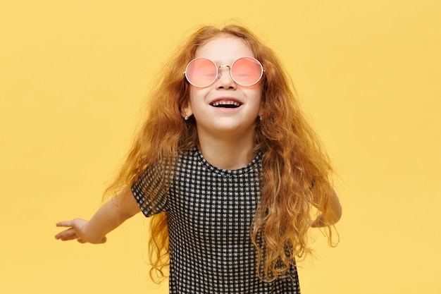 Children, style and fshion concept. Carefree fashionable little girl with curly red hair having happy joyful facial expression, laughing, wearing stylish pink sunglasses, keeping arms behind her back