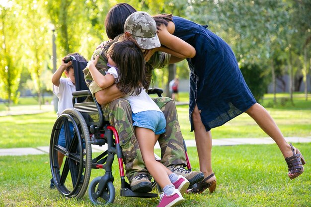 Children and their mom hugging disabled retired military father in park. Veteran of war or returning home concept